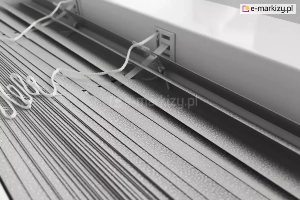 Zdjęcie prezentuje techniczne zbliżenie na produkt żaluzja aluminiowa 16mm w kolorze srebrnym lub jasnoszarym, ukazane z perspektywy górnej. Konstrukcja składa się z bardzo wąskich lameli o szerokości zaledwie szesnastu milimetrów, które charakteryzują się drobną, ziarnistą strukturą powierzchni typu "perforacja" lub "frost", odbijającą światło w sposób rozproszony. Lamele są ciasno ułożone jedna nad drugą, co podkreśla ich smukły profil, idealny do montażu w przestrzeniach z ograniczoną ilością miejsca, np. między listwami przyszybowymi. Centralnym punktem kadru jest miejsce wyjścia białej, plecionej drabinki sznurkowej z rynny górnej, która przechodzi przez precyzyjny, prostokątny otwór w metalowej obudowie. Białe sznurki operacyjne, służące do sterowania mechanizmem, są luźno ułożone na powierzchni zebranych lameli, prezentując swoją teksturę i splot. Tło stanowi fragment białej ramy okiennej lub ściany, co tworzy jasną i sterylną kompozycję kolorystyczną. Całość zdjęcia akcentuje lekkość, precyzję wykonania oraz minimalistyczny design, jaki oferuje żaluzja aluminiowa 16 mm w nowoczesnych systemach osłonowych.