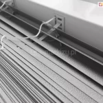 Zdjęcie prezentuje techniczne zbliżenie na produkt żaluzja aluminiowa 16mm w kolorze srebrnym lub jasnoszarym, ukazane z perspektywy górnej. Konstrukcja składa się z bardzo wąskich lameli o szerokości zaledwie szesnastu milimetrów, które charakteryzują się drobną, ziarnistą strukturą powierzchni typu "perforacja" lub "frost", odbijającą światło w sposób rozproszony. Lamele są ciasno ułożone jedna nad drugą, co podkreśla ich smukły profil, idealny do montażu w przestrzeniach z ograniczoną ilością miejsca, np. między listwami przyszybowymi. Centralnym punktem kadru jest miejsce wyjścia białej, plecionej drabinki sznurkowej z rynny górnej, która przechodzi przez precyzyjny, prostokątny otwór w metalowej obudowie. Białe sznurki operacyjne, służące do sterowania mechanizmem, są luźno ułożone na powierzchni zebranych lameli, prezentując swoją teksturę i splot. Tło stanowi fragment białej ramy okiennej lub ściany, co tworzy jasną i sterylną kompozycję kolorystyczną. Całość zdjęcia akcentuje lekkość, precyzję wykonania oraz minimalistyczny design, jaki oferuje żaluzja aluminiowa 16 mm w nowoczesnych systemach osłonowych.