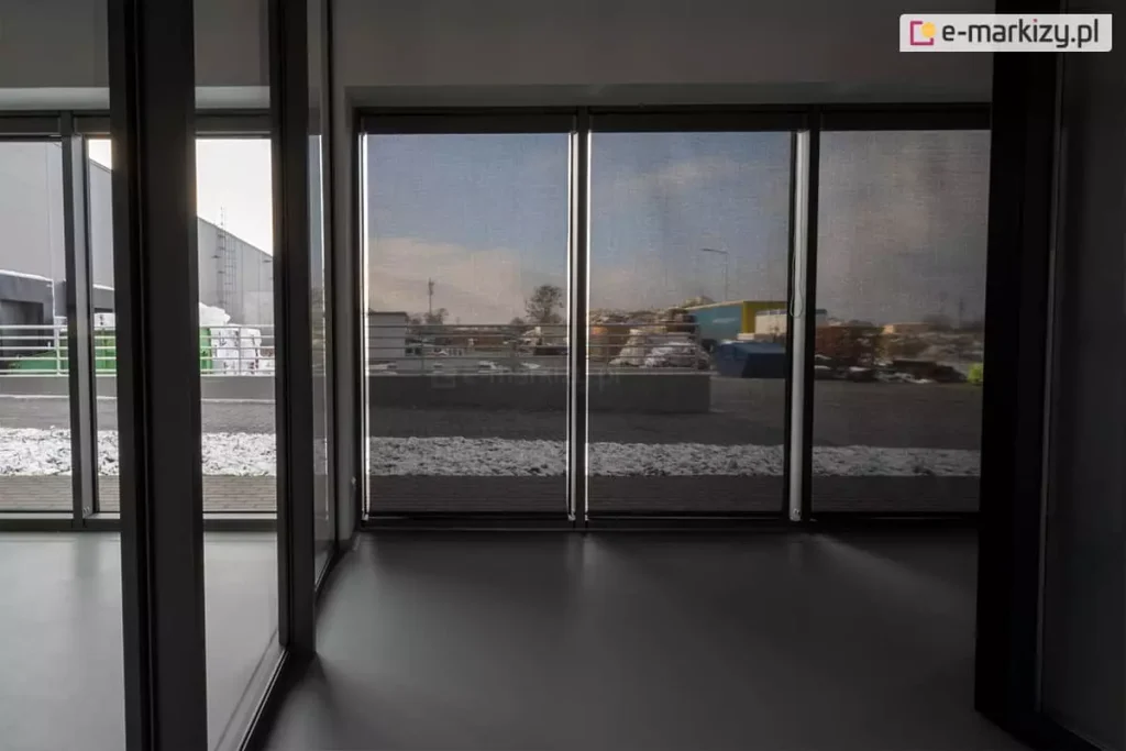Panoramiczne okna zasłonięte roletami screen Aluprof w pomieszczeniu po prawej stronie, prezentujące efekt częściowej przezierności z widocznym zimowym krajobrazem na zewnątrz. Po lewej widoczne wyraźnie lepiej oświetlone pomieszczenie, w którym panoramiczne okna nie zostały przesłonięte refleksolami.