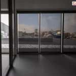 Panoramiczne okna zasłonięte roletami screen Aluprof w pomieszczeniu po prawej stronie, prezentujące efekt częściowej przezierności z widocznym zimowym krajobrazem na zewnątrz. Po lewej widoczne wyraźnie lepiej oświetlone pomieszczenie, w którym panoramiczne okna nie zostały przesłonięte refleksolami.