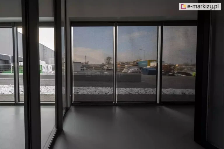 Panoramiczne okna zasłonięte roletami screen Aluprof w pomieszczeniu po prawej stronie, prezentujące efekt częściowej przezierności z widocznym zimowym krajobrazem na zewnątrz. Po lewej widoczne wyraźnie lepiej oświetlone pomieszczenie, w którym panoramiczne okna nie zostały przesłonięte refleksolami.
