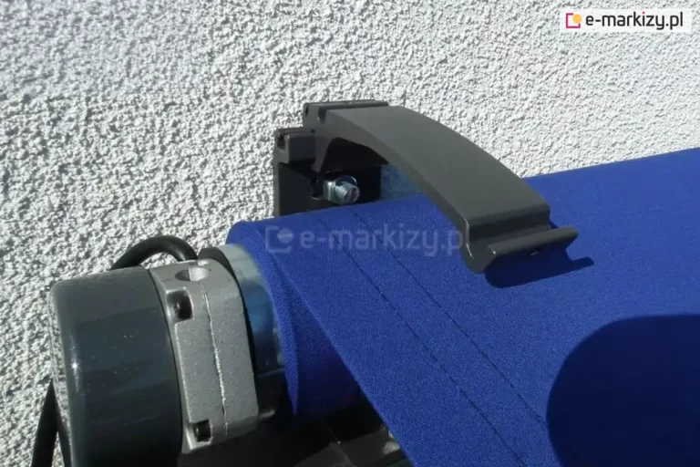 Zbliżenie lewego boku markizy w kolorze konstrukcji srebrzysty grafit z niebieskim poszyciem, widocznym przewodem zasilającym do silnika oraz częściowo wsuniętym w uchwyt ścienny wspornikiem do daszka. W tle ściana z białym tynkiem strukturalnym. Zdjęcie prezentuje montaż daszka do markiz 22,5 cm z mocowaniem ściennym.