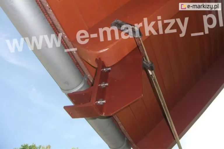 Zbliżenie na techniczny aspekt montażu uchwytów do markizy tarasowej do krokwi dachowej przy użyciu specjalistycznego uchwytu krokwiowego. Solidny aluminiowy wspornik w kolorze brązowym jest solidnie przymocowany do bocznej powierzchni drewnianej belki, która stanowi element podbitki dachu. Śruby mocujące znajdują się po niewidocznej stronie krokwi, a na etapie montażu zostały one dodatkowo wsparte przez zastosowanie zacisków. Uchwyt do krokwi połączony jest z uchwytem ściennym. Mocowanie odbywa się za pomocą trzech śrub przechodzących przez pionową płytę uchwytu, zabezpieczonych nakrętkami. Sam uchwyt ścienny ma formę wygiętej blachy z wystającymi profilami w kształcie litery U, przygotowanymi do osadzenia w nich belki nośnej lub rury markizy. Dodatkowo na zdjęciu widać, że wzdłuż krawędzi dachu przebiega szara, metalowa rynna o przekroju półkolistym, zamontowana tuż nad wspornikiem. W tle widoczne jest pogodne, błękitne niebo oraz wierzchołki drzew w dolnej części kadru.