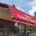Zawieszona nad szeroką witryna sklepową markiza Allegro MOL o łamanym wysięgu z dodatkowym daszkiem osłonowym o białym kolorze. Na czerwonej tkaninie poszycia nadruk "BONBON CUKIERNIA-KAWIARNIA", na falowanej falbanie kolejny nadruk reklamowy "ŚNIADANIA WYPIEKI DOMOWE PRDUKTY EKOLOGICZNE SOKI KAWA". W tle za witryna widoczny niski ceglany murek oraz wysokie iglaki, a nad nimi błękitne niebo z kłębiastymi chmurami nad horyzontem.