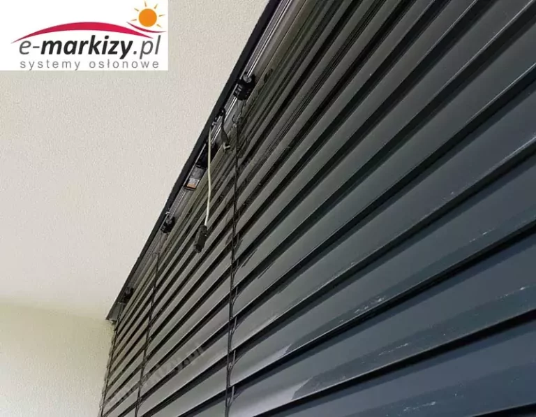Zbliżenie na górną część zewnętrznej żaluzji fasadowej typu C, wykonanej z profili aluminiowych w kolorze antracytowym. Widok od dołu ukazuje szczegóły konstrukcyjne i montażowe systemu osłonowego. Lamele o przekroju w kształcie litery „C” są ustawione w pozycji całkowicie zamkniętej, tworząc jednolitą, zwartą barierę przeciwsłoneczną, chroniącą wnętrze przed nagrzewaniem i zapewniającą pełną prywatność. Na górnej krawędzi żaluzji widoczne są uchwyty montażowe oraz prowadnice taśmowe, przez które przechodzą stalowe taśmy nośne (drabinki) i linki służące do sterowania nachyleniem lamel. W konstrukcji zastosowano system bocznego prowadzenia na taśmach kevlarowych, które cechują się wysoką odpornością na rozciąganie i warunki atmosferyczne. W górnej części kadru widoczna jest również aluminiowa belka górna – element nośny, w którym znajduje się napęd (najczęściej silnik elektryczny) oraz mechanizm odpowiedzialny za podnoszenie i obracanie lamel. Całość systemu została zamontowana w sposób ukryty pod warstwą elewacji, co wskazuje na zastosowanie wariantu podtynkowego, estetycznego i często stosowanego w nowoczesnym budownictwie. Powierzchnia lamel ma delikatny połysk i gładkie wykończenie, co ułatwia ich czyszczenie oraz zwiększa odporność na zabrudzenia i działanie promieni UV. Zdjęcie wykonano pod dużym kątem z perspektywy zewnętrznej, dzięki czemu dobrze widoczna jest symetryczna konstrukcja systemu i sposób jego integracji z elewacją budynku.