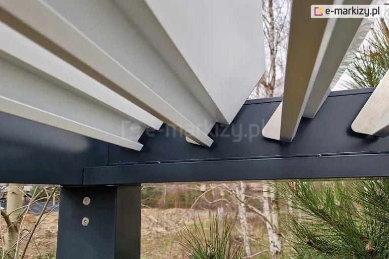 Techniczne zbliżenie na wewnętrzny mechanizm obrotowy lameli pergoli bioklimatycznej SB350 Aluprof. Zdjęcie ukazuje punkt styku białych lameli aluminiowych z boczną szyną prowadzącą w kolorze antracytowym. Lamele są w pozycji otwartej, prezentując srebrne, metalowe zakończenia oraz system trzpieni mocujących. Na pionowym słupie konstrukcyjnym widoczne są dwa łby śrub montażowych typu inbus. W tle, pod konstrukcją, widoczny jest fragment ogrodu z gałęziami sosny, brzozami oraz zarysem budynków mieszkalnych w oddali. Fotografia precyzyjnie dokumentuje spasowanie elementów ruchomych z ramą nośną pergoli.