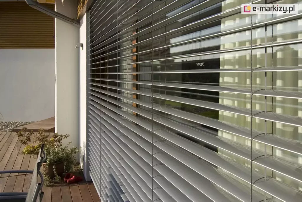 Widok na żaluzje fasadowe C80 zamontowane na elewacji nowoczesnego domu jednorodzinnego. System składa się z licznych, poziomych lameli aluminiowych w kolorze srebrnym, które są ustawione w pozycji otwartej, umożliwiając wgląd do wnętrza przez duże przeszklenie tarasowe. Konstrukcja jest stabilizowana pionowo przez system drabinek i taśm, przechodzących przez każde pióro żaluzji. Po lewej stronie widoczny jest fragment białej, otynkowanej ściany z zewnętrzną oprawą oświetleniową oraz rura spustowa systemu odwodnienia dachu. Żaluzje osłaniają wyjście na drewniany taras, na którym znajduje się donica z roślinnością oraz fragment mebla ogrodowego. W szybach odbija się zieleń ogrodu oraz sąsiednia zabudowa, co podkreśla integrację systemu osłonowego z architekturą budynku i jego otoczeniem.