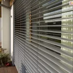 Widok na żaluzje fasadowe C80 zamontowane na elewacji nowoczesnego domu jednorodzinnego. System składa się z licznych, poziomych lameli aluminiowych w kolorze srebrnym, które są ustawione w pozycji otwartej, umożliwiając wgląd do wnętrza przez duże przeszklenie tarasowe. Konstrukcja jest stabilizowana pionowo przez system drabinek i taśm, przechodzących przez każde pióro żaluzji. Po lewej stronie widoczny jest fragment białej, otynkowanej ściany z zewnętrzną oprawą oświetleniową oraz rura spustowa systemu odwodnienia dachu. Żaluzje osłaniają wyjście na drewniany taras, na którym znajduje się donica z roślinnością oraz fragment mebla ogrodowego. W szybach odbija się zieleń ogrodu oraz sąsiednia zabudowa, co podkreśla integrację systemu osłonowego z architekturą budynku i jego otoczeniem.
