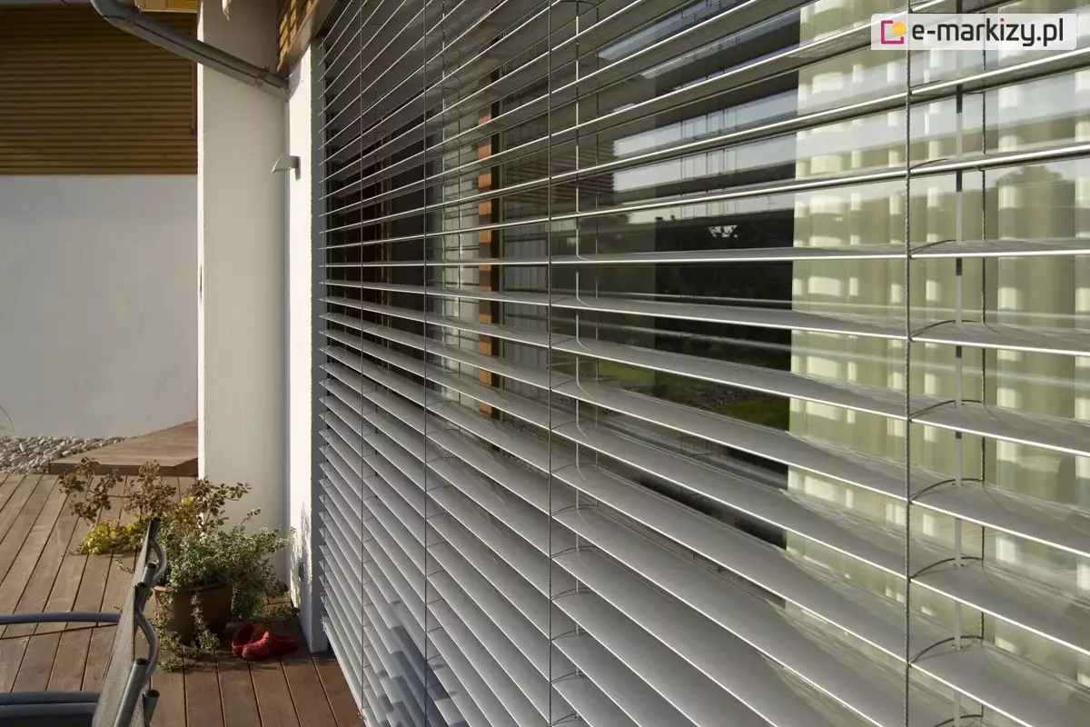 Widok na żaluzje fasadowe C80 zamontowane na elewacji nowoczesnego domu jednorodzinnego. System składa się z licznych, poziomych lameli aluminiowych w kolorze srebrnym, które są ustawione w pozycji otwartej, umożliwiając wgląd do wnętrza przez duże przeszklenie tarasowe. Konstrukcja jest stabilizowana pionowo przez system drabinek i taśm, przechodzących przez każde pióro żaluzji. Po lewej stronie widoczny jest fragment białej, otynkowanej ściany z zewnętrzną oprawą oświetleniową oraz rura spustowa systemu odwodnienia dachu. Żaluzje osłaniają wyjście na drewniany taras, na którym znajduje się donica z roślinnością oraz fragment mebla ogrodowego. W szybach odbija się zieleń ogrodu oraz sąsiednia zabudowa, co podkreśla integrację systemu osłonowego z architekturą budynku i jego otoczeniem.