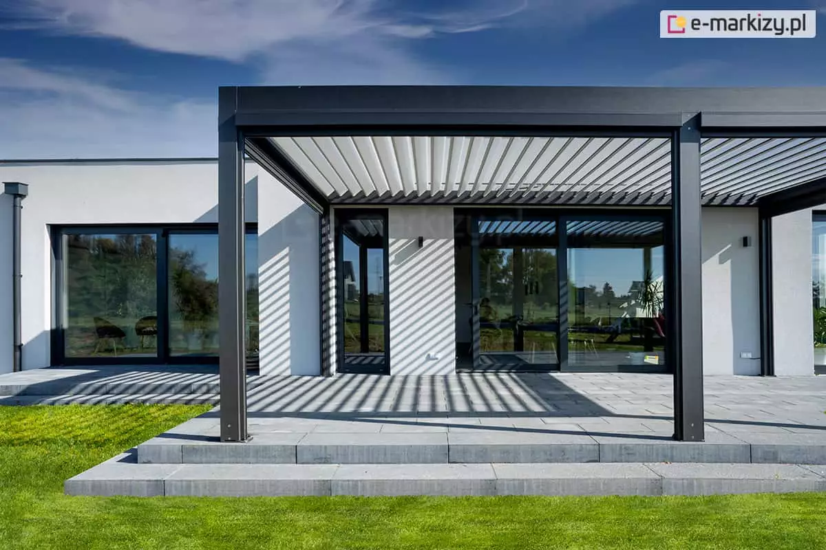 Opcjonalna podpora środkowa może okazać się niezbędna do montażu refleksoli, czyli dodatkowych osłon bocznych. Pergola SB400 w dwóch kolorach współgra z kolorystyką otoczenia oraz nowoczesną bryłą parterowego budynku o jasnej elewacji i ciemnych ramach dużych przeszkleń.