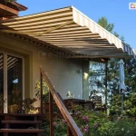 Zdjęcie prezentuje w pełni rozłożoną markizę Piano, zamontowaną na bocznej ścianie tradycyjnego domu z ogrodem. Poszycie markizy charakteryzuje się klasycznym wzorem w szerokie, poziome pasy w kolorach piaskowego beżu i bieli. Tkanina jest precyzyjnie napięta na aluminiowych ramionach przegubowych o białym wykończeniu, które harmonizuje z kolorystyką poszycia. Przednia krawędź markizy Piano została zakończona ozdobną, falistą falbaną z białą lamówką. Konstrukcja jest zamontowana na beżowej elewacji budynku, tuż obok drewnianych schodów prowadzących na podwyższony taras. Wokół tarasu widoczna jest bujna roślinność, w tym kwitnące na różowo krzewy oraz wysokie drzewa iglaste, które tworzą naturalne tło. Na samym tarasie znajdują się drewniane meble ogrodowe oraz złożony parasol przeciwsłoneczny. Mocne światło słoneczne pada na powierzchnię markizy Piano, podkreślając jej funkcję ochronną i tworząc wyraźną strefę cienia nad wejściem do domu.