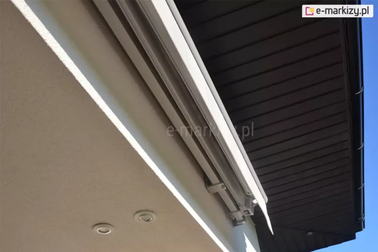 Zamocowana na ścianie pod podbitką dachu w ciemnym kolorze markiza 4 metrowa Jamaica o białej konstrukcji i jasnej tkaninie. W suficie pod ścianą z markizą widoczne dwa punkty oświetlenia.