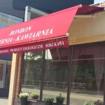 Zdjęcie prezentuje zastosowanie komercyjne produktu, jakim jest markiza tarasowa Allegro, zamontowana nad witryną cukierni i kawiarni Bonbon. Intensywnie czerwone poszycie markizy pełni funkcję zarówno ochronną, jak i reklamową, posiadając naniesione duże białe napisy z nazwą lokalu oraz listą oferowanych produktów, takich jak śniadania, wypieki domowe, produkty ekologiczne, soki i kawa. Markiza jest w pełni rozwinięta, a jej przednia część opada pod kątem, tworząc szeroki daszek osłaniający przeszkloną witrynę sklepową przed bezpośrednim nasłonecznieniem. Dolna krawędź tkaniny wykończona jest dekoracyjną, falowaną falbaną, na której również widnieją białe napisy informacyjne. Konstrukcja markizy, w tym ramiona przegubowe i belka montażowa, jest polakierowana na kolor biały, co estetycznie komponuje się z jasną elewacją budynku oraz ceglanymi filarami wokół okien. Pod markizą, na parapecie kawiarni, ustawione są donice z białymi kwiatami, a w szybach odbija się ulica i sąsiednia zabudowa. Całość ukazuje, jak markiza tarasowa Allegro może efektywnie wspierać identyfikację wizualną punktu usługowego, jednocześnie poprawiając komfort klientów wewnątrz lokalu.