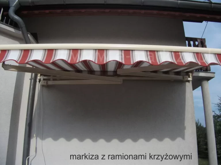 Zdjęcie prezentuje specjalistyczny model, jakim jest markiza Australia z ramionami krzyżowymi, zainstalowana na szarej, tynkowanej elewacji budynku. Charakterystyczną cechą tej konstrukcji jest zastosowanie ramion o różnej wysokości montażu, co pozwala im na krzyżowanie się podczas składania i rozwijania, umożliwiając uzyskanie wysięgu większego niż szerokość samej markizy. Ramiona przegubowe oraz rura osłonowa wykonane są z aluminium lakierowanego na kolor kremowy. Poszycie markizy stanowi pasiaste płótno w odcieniach intensywnej czerwieni, bieli oraz szarości, zakończone ozdobną falbaną o falistym brzegu. Markiza Australia z ramionami krzyżowymi jest częściowo rozwinięta, rzucając na ścianę budynku wyraźny cień o ząbkowanym profilu, zgodnym z kształtem falbany. Powyżej markizy widoczny jest fragment dachu z ciemną rynną oraz balkon z balustradą, a po prawej stronie konstrukcji znajduje się betonowy filar podtrzymujący górną kondygnację. Po lewej stronie zdjęcia biegnie pionowa rura spustowa oraz biały przewód instalacyjny. Tło stanowi błękitne niebo oraz wierzchołki drzew iglastych, co sugeruje montaż w budynku mieszkalnym lub rekreacyjnym.