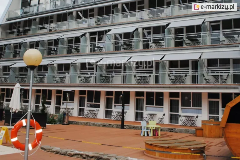 Zdjęcie przedstawia szerokie ujęcie wielopiętrowego budynku hotelowego lub pensjonatu, na którego balkonach zainstalowano liczne markizy balkonowe. Budynek posiada nowoczesną architekturę z białymi elewacjami i szklanymi balustradami wykończonymi metalowymi poręczami. Na trzech kondygnacjach widoczne są powtarzalne moduły balkonowe wyposażone w jasnoszare markizy. Osłony te są zamontowane pod sufitami wyższych pięter i oparte na bocznych ramionach, tworząc jednolity pas ochrony przeciwsłonecznej wzdłuż całej fasady. Na każdym balkonie znajdują się zestawy mebli wypoczynkowych składające się z okrągłego, ciemnego stolika oraz dwóch ażurowych krzeseł. Parter budynku posiada otwarte wyjścia na przestronny taras wyłożony deską kompozytową w kolorze jasnobrązowym. Na tarasie ustawiono stoliki kawiarniane, parasole ogrodowe oraz niewielki zestaw mebli dziecięcych z kolorowymi krzesełkami. W lewym dolnym rogu zdjęcia widoczny jest element infrastruktury basenowej lub rekreacyjnej – pomarańczowe koło ratunkowe zawieszone na metalowym słupku z lampą o kulistym kloszu. Po prawej stronie kadru znajduje się fragment drewnianej konstrukcji przypominającej beczkę lub saunę zewnętrzną. Całość ujęcia prezentuje kompleksowe zastosowanie systemów osłonowych w obiekcie komercyjnym, dbając o komfort użytkowników na wielu poziomach jednocześnie.