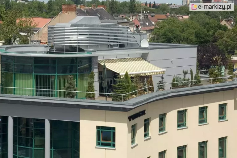 Widok z lotu ptaka na rozległy taras umieszczony na dachu nowoczesnego budynku biurowego lub apartamentowca. Na tarasie rozłożona jest duża osłona przeciwsłoneczna - markiza Palladio Aluprof o poszyciu w jasne, żółto-białe pionowe pasy, wykończona dekoracyjną falbaną na krawędzi. Konstrukcja osłania strefę wypoczynkową, w której ustawione są liczne rośliny iglaste w donicach, tworzące zielony szpaler wzdłuż metalowej barierki zabezpieczającej krawędź dachu. Budynek posiada elewację w kolorze piaskowym z licznymi oknami w zielonych ramach oraz nowoczesną, przeszkloną nadbudówkę o półkolistym kształcie. Na dachu widoczne są również elementy infrastruktury technicznej, w tym srebrne osłony wentylacyjne o lamelowej budowie oraz antena satelitarna. W tle rozpościera się panorama miasta z widocznymi dachami kamienic, zielonymi koronami drzew oraz jasnym, lekko zamglonym niebem.