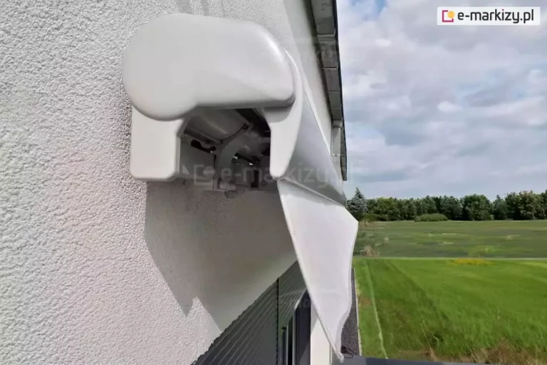 Zawieszona na białej ścianie nad oknami przesłoniętymi ciemnymi roletami zewnętrznymi biała markiza Palladio z białym poszyciem widocznym w postaci prostej falbany, widoczna od boku. Na pierwszym planie widoczna dopasowana kolorystycznie do aluminiowej kasety plastikowa zaślepka boczna. Ponad markizą widoczny system rynnowy budynku. Po jej prawej stronie rozciąga się rozległy widok na zielone łąki oraz drzewa na horyzoncie, a nad nimi lekko szarawe przychmurzone niebo.