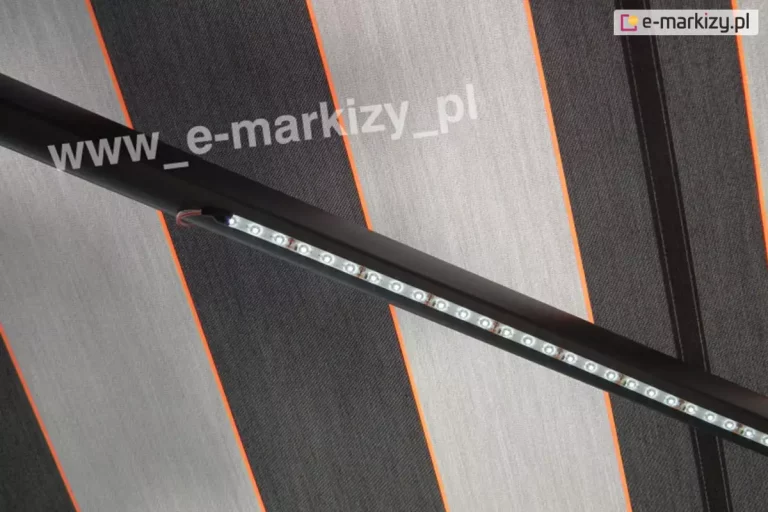 Zbliżenie ramienia markizy z zamocowanym do niej oświetleniem LED do markiz o barwie zimnej. Ramię w kolorze srebrzysty grafit z taśmą led widoczne jest na tle tkaniny poszycia w szerokie pasy szare i grafitowe oraz wąskie pasy czerwone pomiędzy nimi.