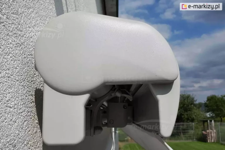 Markiza Palladio Aluprof w całości złożona widoczna od lewego boku. Pod plastikową zaślepką boczną widoczny wspornik kasety oraz złożone ramię tuż przy belce przedniej, w której widoczny skrawek powiewającej na wietrze falbany. Markiza zamocowana na białej ścianie, obok niej widoczny ogród oraz drzewa z zielonymi liśćmi i zachmurzone niebo.