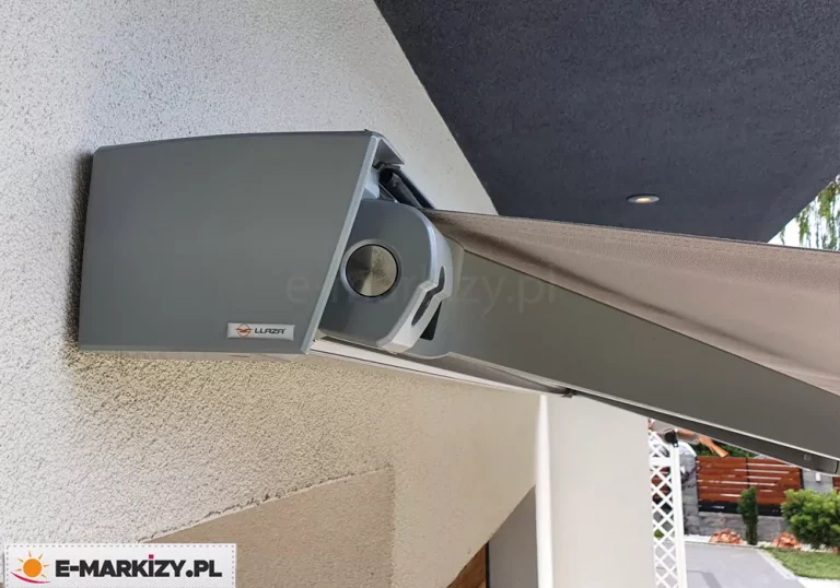 markiza automatyczna moderno MOL z charakterystyczną kwadratową kasetą w kolorze srebrnym, zamocowana do ściany nad wnęką i pod podświetlonym sufitem tarasowym. W tle widoczny płot i zielone krzewy.