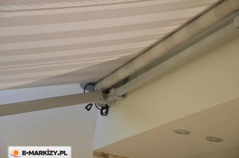 Zbliżenie na mechanizm konstrukcyjny oraz materiał poszycia markiza Jamaica zamontowanej do ściany budynku. Zdjęcie prezentuje detale techniczne lewej strony markizy, gdzie widoczna jest aluminiowa rura nawojowa z nawiniętą tkaniną oraz ramię przegubowe odpowiedzialne za rozkładanie i napinanie materiału. Tkanina markizowa posiada wzór w szerokie pasy w odcieniach bieli i jasnego beżu, a jej powierzchnia jest lekko pofalowana pod wpływem naprężenia. Cała konstrukcja markiza Jamaica, w tym profil nośny, ramię oraz rura, polakierowana jest proszkowo na kolor biały o matowym wykończeniu. Pod rurą nawojową widoczne są czarne przewody elektryczne doprowadzone do silnika sterującego, które są estetycznie wyprowadzone z mechanizmu. Markiza jest przytwierdzona do jasnej elewacji wykończonej tynkiem strukturalnym, a poniżej punktu montażu znajduje się fragment wnęki sufitowej z dwoma wpuszczanymi punktami oświetleniowymi w srebrnych oprawach. Kąt nachylenia ramienia sugeruje, że osłona jest w trakcie rozkładania lub znajduje się w pozycji częściowego wysięgu. Wszystkie elementy metalowe, takie jak śruby montażowe i przeguby, są precyzyjnie dopasowane, co podkreśla solidność i techniczne zaawansowanie modelu markiza Jamaica.