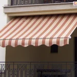 Zdjęcie prezentuje montaż markizy Piano MOL do sufitu. Na obrazie przedstawiono klasyczną markizę Piano na balkon, zamontowaną we wnęce loggii nowoczesnego, białego budynku mieszkalnego. Poszycie markizy wykonane jest z trwałej tkaniny w szerokie, pionowe pasy w kolorach ceglanego brązu oraz jasnego beżu, co tworzy elegancki kontrast z jasną elewacją. Materiał jest w pełni rozwinięty i napięty na konstrukcji, skutecznie osłaniając przestrzeń balkonu przed słońcem. Przednia krawędź, jaką posiada markiza Piano na balkon, wykończona jest ozdobną falbaną o falistym kształcie, obszytą lamówką dopasowaną kolorystycznie do ciemniejszych pasów tkaniny. Aluminiowy profil frontowy oraz widoczne zaślepki boczne są polakierowane na czysty, biały kolor. Poniżej linii markizy widoczna jest ozdobna, czarna balustrada balkonowa o geometrycznym wzorze, a powyżej znajduje się fragment kolejnego balkonu z podobną barierką. Markiza Piano na balkon została zainstalowana pod stropem wyższej kondygnacji, co zapewnia stabilność montażu i estetyczne domknięcie wnęki balkonowej.