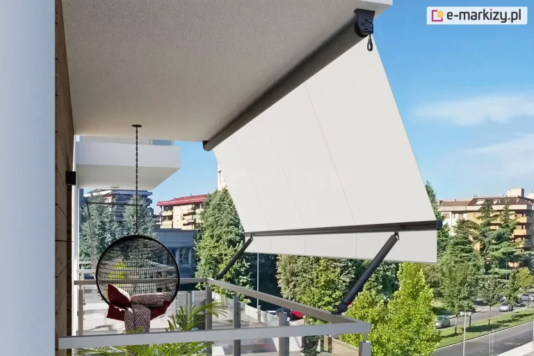 Widok z boku na nowoczesny balkon wyposażony w markizę balkonową z ramionami poręczowymi, zamontowaną pod sufitem wnęki. Markiza posiada poszycie z gładkiej tkaniny w kolorze kremowym lub bardzo jasnym beżu, która jest rozwinięta i napięta pod kątem, wychodząc poza obrys barierki. Konstrukcja wspiera się na dwóch ciemnych, antracytowych ramionach bocznych przymocowanych do balustrady, które utrzymują dolną belkę dociążającą. Na suficie widoczna jest rura nawojowa z mechanizmem korbowym zakończonym oczkiem. Na tarasie, w cieniu osłony, znajduje się wiszący fotel typu kokon o czarnym, ażurowym splotcie, zawieszony na metalowym łańcuchu, wyłożony wzorzystymi poduszkami. Obok fotela widać fragmenty zielonych roślin doniczkowych. Balustrada balkonu jest minimalistyczna, wykonana z jasnych profili metalowych z wypełnieniem szklanym. W tle rozpościera się widok na miejską panoramę z wielopiętrowymi budynkami mieszkalnymi, gęstą zielenią drzew oraz fragmentem ulicy z zaparkowanymi samochodami pod bezchmurnym, błękitnym niebem.