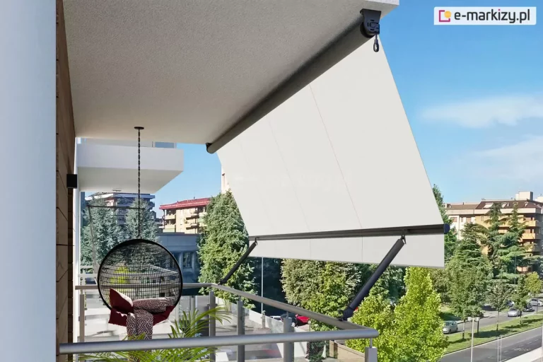 Widok z boku na nowoczesny balkon wyposażony w markizę balkonową z ramionami poręczowymi, zamontowaną pod sufitem wnęki. Markiza posiada poszycie z gładkiej tkaniny w kolorze kremowym lub bardzo jasnym beżu, która jest rozwinięta i napięta pod kątem, wychodząc poza obrys barierki. Konstrukcja wspiera się na dwóch ciemnych, antracytowych ramionach bocznych przymocowanych do okrągłej balustrady, które utrzymują dolną belkę dociążającą. Na suficie widoczna jest rura nawojowa z mechanizmem korbowym zakończonym oczkiem. Na tarasie, w cieniu osłony, znajduje się wiszący fotel typu kokon o czarnym, ażurowym splocie, zawieszony na metalowym łańcuchu, wyłożony wzorzystymi poduszkami. Obok fotela widać fragmenty zielonych roślin doniczkowych. Balustrada balkonu jest minimalistyczna, wykonana z jasnych profili metalowych z wypełnieniem szklanym. W tle rozpościera się widok na miejską panoramę z wielopiętrowymi budynkami mieszkalnymi, gęstą zielenią drzew oraz fragmentem ulicy z zaparkowanymi samochodami pod bezchmurnym, błękitnym niebem.
