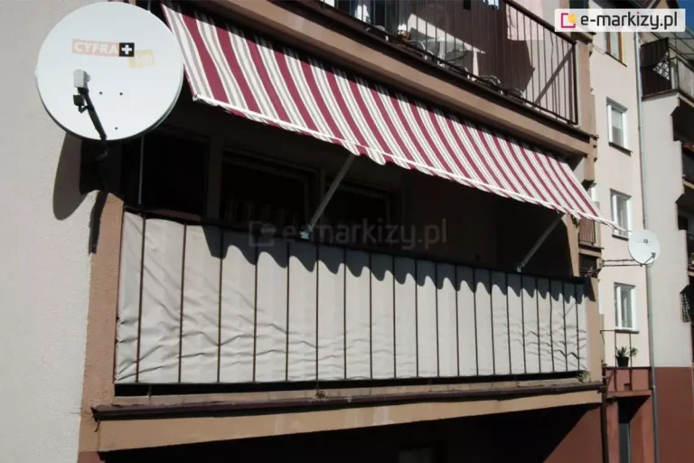Zdjęcie przedstawia szeroki widok na balkon w bloku mieszkalnym, na którym zainstalowano markizę balkonową oraz osłonę balustrady. Markiza posiada poszycie w klasyczne pionowe pasy w kolorze bordowym i białym (kremowym), a jej dolna krawędź wykończona jest falbaną o falistym kształcie z dopasowaną lamówką. Konstrukcja markizy opiera się na dwóch białych ramionach bocznych, które są zamocowane do balustrady, pozwalając na nachylenie materiału pod kątem. Poniżej, na całej długości balustrady, zamontowana jest techniczna osłona przeciwwiatrowa i widokowa w kolorze beżowym, przepleciona między pionowymi szczeblami barierek. Po lewej stronie balkonu, na elewacji budynku, zamontowana jest biała antena satelitarna z logotypem operatora. Budynek posiada jasną elewację z brązowymi akcentami wokół wnęki balkonowej. W tle widoczne są sąsiednie balkony oraz fragmenty innych budynków wielorodzinnych, co wskazuje na typową zabudowę miejską. Całość ukazuje praktyczne wykorzystanie osłon zewnętrznych w celu zwiększenia prywatności i ochrony przed słońcem na balkonie w bloku.