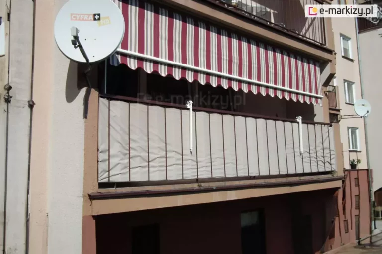 Zdjęcie przedstawia zbliżenie na balkon w bloku mieszkalnym, na którym zainstalowana została markiza balkonowa poręczowa. Poszycie markizy wykonane jest z materiału o wzorze w pionowe pasy w kolorach bordowym, szarym i kremowym. Markiza jest częściowo rozwinięta, a jej dolna krawędź kończy się dekoracyjną, falistą falbaną z białą lamówką. Biała rura przednia markizy opiera się na dwóch pionowych ramionach mocowanych do poręczy, co pozwala na stabilne ustawienie kąta nachylenia tkaniny. Balustrada balkonu, wykonana z ciemnych, metalowych profili, jest niemal w całości zasłonięta gęstą, beżową matą osłonową, która zapewnia prywatność i chroni przed wiatrem. Po lewej stronie, na elewacji budynku, widoczna jest biała, okrągła antena satelitarna z logotypem operatora cyfrowego, zamontowana na ciemnym wysięgniku. Ściana budynku wykończona jest tynkiem w kolorze jasnopiaskowym, natomiast dolna część balkonu i pas pod nim mają ciemniejszy, czerwonobrązowy odcień. W tle, po prawej stronie, widać fragmenty sąsiednich balkonów oraz elewację dalszej części bloku z oknami. Całość ujęcia charakteryzuje się ostrym, dziennym światłem, które rzuca wyraźny cień markizy na wnękę balkonową.