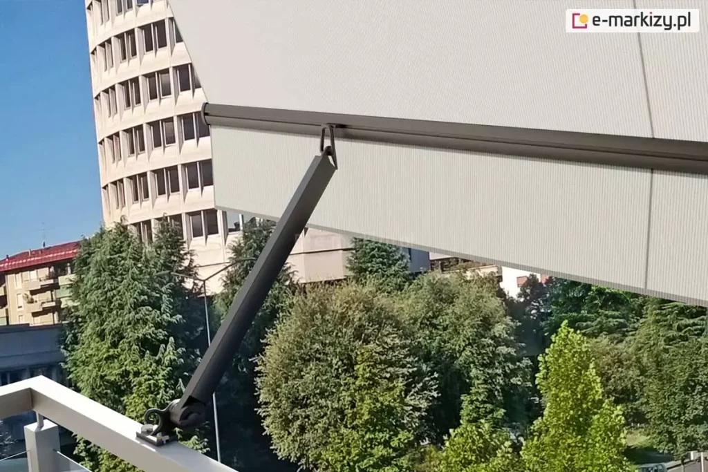 Ramię markizy balkonowej Italia poręczowa w kolorze srebrzysty grafit zamocowane do jasnej barierki metalowej balustrady balkonu. W uniesionym ramieniu zaczepiona została belka przednia, z której zwisa prosta falbana oraz napięte jest poszycie. Pod falbaną widoczne liczne drzewa o zielonych liściach. Z lewej strony za markizą widoczna zaokrąglona fasada wysokiego budynku o wielu oknach oraz niższe sąsiednie bloki mieszkalne.