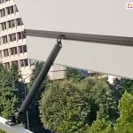 Ramię markizy balkonowej Italia poręczowa w kolorze srebrzysty grafit zamocowane do jasnej barierki metalowej balustrady balkonu. W uniesionym ramieniu zaczepiona została belka przednia, z której zwisa prosta falbana oraz napięte jest poszycie. Pod falbaną widoczne liczne drzewa o zielonych liściach. Z lewej strony za markizą widoczna zaokrąglona fasada wysokiego budynku o wielu oknach oraz niższe sąsiednie bloki mieszkalne.