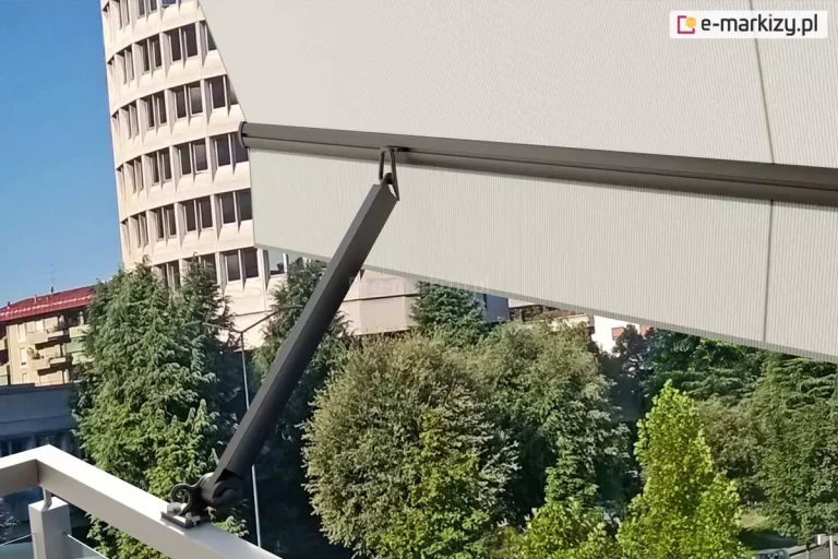 Ramię markizy balkonowej Italia poręczowa w kolorze srebrzysty grafit zamocowane do jasnej barierki metalowej balustrady balkonu. W uniesionym ramieniu zaczepiona została belka przednia, z której zwisa prosta falbana oraz napięte jest poszycie. Pod falbaną widoczne liczne drzewa o zielonych liściach. Z lewej strony za markizą widoczna zaokrąglona fasada wysokiego budynku o wielu oknach oraz niższe sąsiednie bloki mieszkalne.