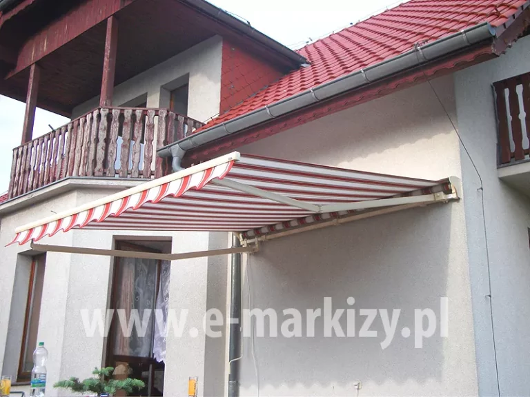 Markiza w biało czerwone pasy z falowaną falbaną i białą konstrukcją zawieszona na jasnej elewacji piętrowego budynku z balkonem i dachem krytym czerwoną dachówką. markiza zamocowana przy narożniku budynku w pobliżu okien w wykuszu.