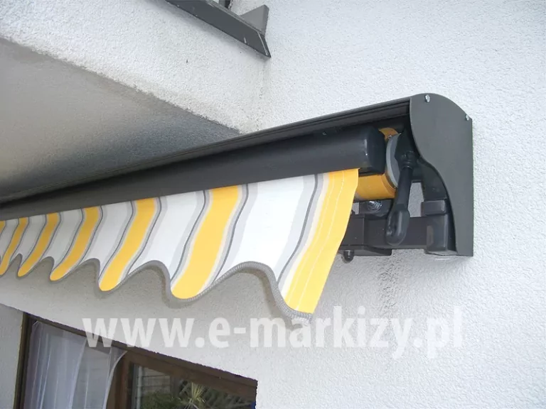 Markiza bez kasety z opcjonalnym daszkiem do markiz w kolorze dopasowanym do barwy konstrukcji z tkaniną falbany w białe, żółte i szare pasy, wyciętą w kształt fali, zamocowana na ścianie z jasnym tynkiem strukturalnym pod balkonem i nad oknem tarasowym. Po prawej stronie markizy pod daszkiem widoczna przekładnia do napędu ręcznego korbowego.