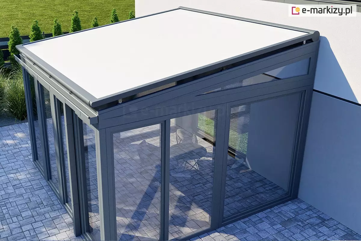 nowoczesny ogród zimowy o konstrukcji aluminiowo-szklanej, widziany z góry pod lekkim kątem, od strony zewnętrznego narożnika. Na szklanym dachu obiektu zainstalowano zewnętrzny system Veranda V-Ziiip selt z jasnym, białym poszyciem, które jest w pełni rozwinięte i precyzyjnie napięte. Konstrukcja nośna ogrodu zimowego, w tym ramy okienne, profile dachowe oraz prowadnice verandy V-Ziiip, są polakierowane na elegancki, matowy kolor antracytowy. Ściany boczne i frontowe obiektu są w całości przeszklone, co pozwala dostrzec wnętrze z jasną, brukowaną posadzką oraz zarysem mebli wypoczynkowych. Ogród zimowy przylega do jasnej elewacji budynku, a wokół niego rozciąga się starannie utrzymany trawnik oraz rząd tui wzdłuż ogrodzenia w tle. Perspektywa zdjęcia uwypukla techniczne aspekty montażu osłony przeciwsłonecznej oraz nowoczesny design całej bryły architektonicznej, łączącej ciemne profile z jasnym materiałem markizy.