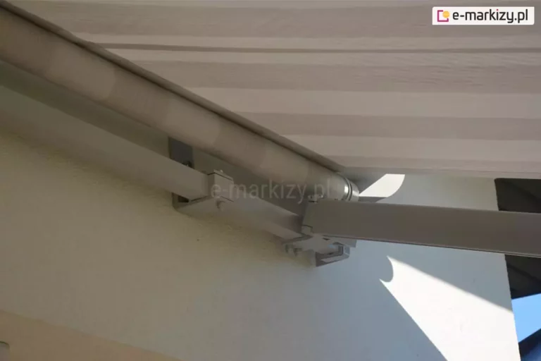 Zdjęcie prezentuje zbliżenie na prawą stronę mechanizmu markizy Jamaica, ukazując solidny system mocowania oraz ramię przegubowe w pozycji rozłożonej. Aluminiowa konstrukcja markizy, w tym belka kwadratowa nośna oraz ramię, została polakierowana proszkowo na kolor biały. Widoczny jest boczny uchwyt ramienia, który łączy się z belką nośną za pomocą stalowych śrub, zapewniając stabilność całego systemu. Tkanina markizowa w szerokie, poziome pasy w kolorze białym i jasnobeżowym jest nawinięta na stalową rurę nawojową, która znajduje się bezpośrednio nad ramieniem. Markiza Jamaica została zamontowana do jasnej, otynkowanej elewacji budynku. W prawym dolnym rogu zdjęcia widać krawędź ściany, za którą przebiega linia dachu z ciemną podbitką. Oświetlenie naturalne pada z prawej strony, rzucając delikatny cień konstrukcji na białą ścianę, co podkreśla trójwymiarowość i masywność elementów mechanicznych. Detale takie jak wykończenie krawędzi profilu aluminiowego oraz precyzyjne ściegi na materiale świadczą o wysokim standardzie wykonania markizy Jamaica Aluprof