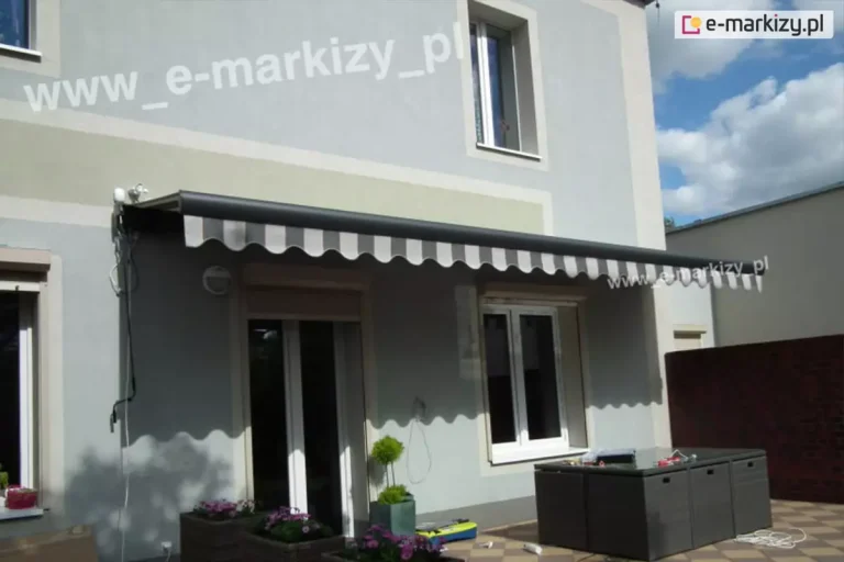 Zdjęcie prezentuje nowoczesny budynek piętrowy o szarej elewacji, na którym zainstalowana została markiza elektryczna z awaryjnym otwieraniem model Palladio Aluprof. Markiza jest częściowo rozwinięta, a jej poszycie wykonane jest z wytrzymałej tkaniny w szerokie, pionowe pasy w odcieniach szarości i bieli, co idealnie komponuje się z kolorystyką fasady. Przedni profil markizy jest w kolorze ciemnoantracytowym i posiada ozdobną, pofalowaną falbanę, która rzuca charakterystyczny, ząbkowany cień na ścianę budynku. Konstrukcja została zamontowana nad wyjściem tarasowym i oknem parteru, osłaniając przestrzeń wypoczynkową. Na lewym boku kasety widoczny jest mechanizm napędowy z wyprowadzonym przewodem zasilającym oraz uchem do korby, co wskazuje, że jest to markiza elektryczna z awaryjnym otwieraniem, umożliwiająca ręczne zwinięcie osłony w przypadku braku prądu. Taras przed domem wyłożony jest dwukolorową kostką brukową w układzie szachownicy, a w jego centralnej części znajduje się zestaw nowoczesnych mebli ogrodowych w formie ciemnoszarych modułów. Całość dopełniają detale takie jak donice z kwiatami oraz jasne obramowania wokół okien, a w tle widać błękitne niebo z lekkim zachmurzeniem.