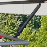 Okrągła barierka balustrady balkonu z metalu z zamocowanym do niej uchwytem ramienia poręczowego w kolorze czarnym z zamocowana w niej belką przednią z tkaniną w kolorze kremowym. Pod prostą falbaną widoczne zielone drzewa liściaste.