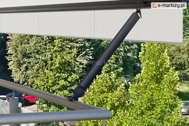 Okrągła barierka balustrady balkonu z metalu z zamocowanym do niej uchwytem ramienia poręczowego w kolorze czarnym z zamocowana w niej belką przednią z tkaniną w kolorze kremowym. Pod prostą falbaną widoczne zielone drzewa liściaste.