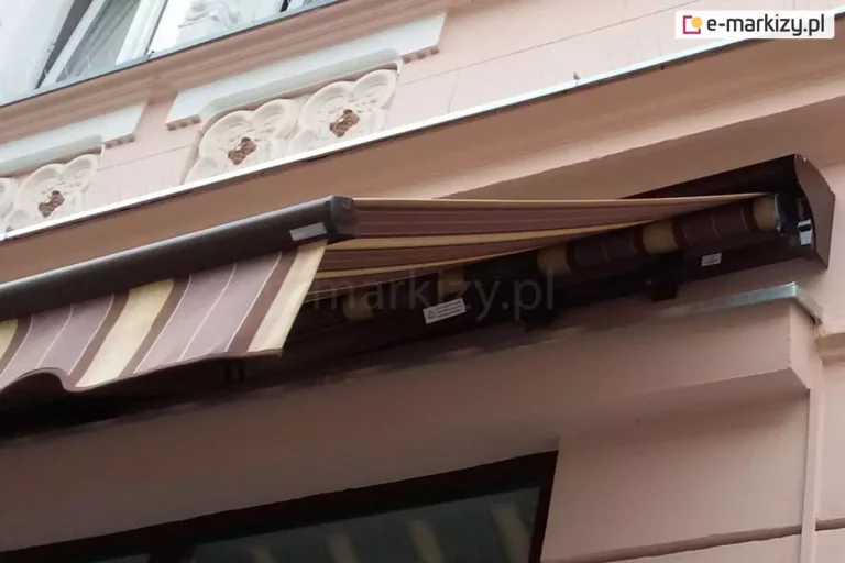 Zdjęcie prezentuje markizę tarasową Jamaica z dodatkową osłoną tkaniny w postaci opcjonalnego daszka, zamontowaną na elewacji zabytkowej kamienicy o bogatych zdobieniach architektonicznych. Konstrukcja markizy, w tym profil przedni oraz boczna pokrywa daszka, wykonana jest z aluminium lakierowanego na kolor ciemnobrązowy. Daszek ma nowoczesny, lekko zaokrąglony kształt, który po całkowitym zwinięciu markizy skutecznie chroni tkaninę przed czynnikami zewnętrznymi. Poszycie markizy wykonane jest z materiału w szerokie pasy w odcieniach brązu i beżu, co komponuje się z kolorystyką profilu. Z przodu widoczna jest ozdobna falbana o falistym brzegu, wykończona lamówką, która zwisa pionowo z profilu przedniego. Markiza jest częściowo rozwinięta, odsłaniając rurę nawojową oraz mechanizm ukryty pod kasetą. Montaż został wykonany nad oknem, pod ozdobnym gzymsem elewacji, która jest utrzymana w kolorze brudnego różu z białymi detalami sztukateryjnymi w formie motywów roślinnych. Zdjęcie wykonane z perspektywy dolnej ukazuje sposób integracji nowoczesnej osłony przeciwsłonecznej z klasyczną fasadą budynku.