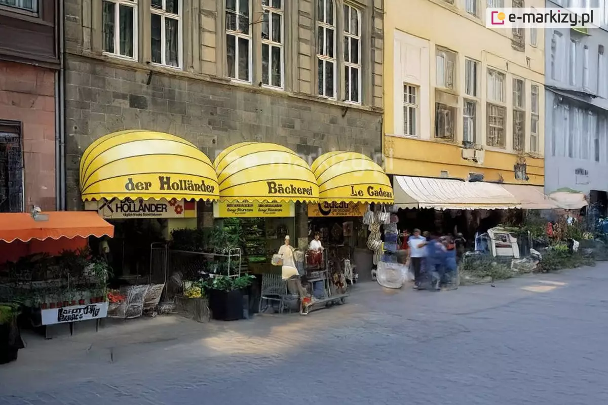 Zdjęcie przedstawia miejską pierzeję z rzędem punktów handlowo-usługowych, nad którymi zamontowano różnorodne markizy, z dominacją trzech dużych markiz koszowych w jaskrawym kolorze. Markizy koszowe mają kształt półkolisty i wykonane są z połyskującego materiału pcv w kolorze intensywnie żółtym, przedzielonego poziomymi, czarnymi liniami podkreślającymi ich konstrukcję. Na każdej z nich znajduje się czarny napis identyfikujący lokal: „der Holländer”, „Bäckerei” oraz „Le Cadeau”. Dolne krawędzie markiz wykończone są falowaną falbaną z czarną lamówką. Pod pierwszą markizą widoczny jest sklep z roślinami i kwiatami, przed którym wystawiono liczne donice. Lokale mieszczą się w zabytkowej kamienicy o szarej, kamiennej elewacji z wysokimi, pionowymi oknami. Po lewej stronie widoczna jest mniejsza, klasyczna markiza tarasowa w kolorze pomarańczowym, natomiast po prawej stronie ciągu handlowego zamontowano jasne markizy o prostym poszyciu. Przed sklepami rozciąga się szeroki, wybrukowany plac lub chodnik, po którym poruszają się przechodnie. Światło słoneczne pada z boku, tworząc wyraźne cienie pod konstrukcjami osłonowymi i podkreślając jaskrawą barwę żółtych poszyć, które stanowią główny akcent kolorystyczny ulicy.
