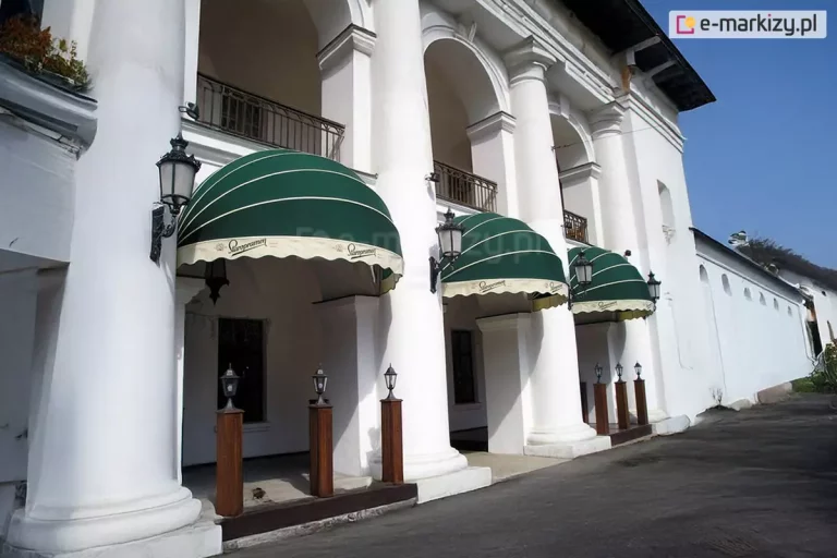 Zdjęcie przedstawia fragment zabytkowej białej budowli z kolumnadą, w której łukach zamontowano trzy eleganckie markizy koszowe o półkolistym kształcie. Poszycie markiz wykonane jest z tkaniny w głębokim, ciemnozielonym kolorze, którą zdobią cienkie, poziome, białe pasy biegnące wzdłuż łuków stelaża. Dolne krawędzie markiz wykończone są prostymi, jasnymi falbanami w kolorze kremowym, na których nadrukowany jest ozdobny, czarny logotyp marki „Staropramen”. Każda z markiz jest precyzyjnie osadzona między masywnymi, białymi kolumnami o klasycznych bazach i kapitelach. Na kolumnach, po bokach markiz, zamocowane są stylizowane czarne latarnie ścienne z przeszklonymi kloszami. Pod zadaszeniami, na podestach, ustawiono mniejsze, stojące lampy ogrodowe na drewnianych słupkach. Budynek posiada liczne arkady i balkony z ozdobnymi, ciemnymi balustradami na wyższej kondygnacji. Przed budynkiem przebiega asfaltowa droga lub plac. Całość scenerii jest oświetlona jasnym, dziennym światłem pod bezchmurnym, błękitnym niebem, co podkreśla kontrast między zielenią osłon a bielą elewacji i nadaje miejscu prestiżowy charakter lokalu gastronomicznego.