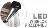 Grafika prezentująca opcję oświetlenia LED w belce przedniej markizy tarasowej. W okrągłym kadrze po lewej stronie widoczne jest ujęcie od dołu profilu czołowego (belki przedniej) w kolorze ciemnego grafitu lub czarnym. Wzdłuż profilu zamontowana jest zintegrowana listwa oświetleniowa, która emituje jasne, białe światło skierowane w dół, oświetlając przestrzeń pod markizą. Powyżej profilu widoczny jest fragment rozwiniętego poszycia materiałowego w brązowo-beżowe pasy. Po prawej stronie grafiki znajduje się duży, stylizowany napis „LED” ułożony z punktów przypominających diody, a poniżej tekst drukowanymi literami „W BELCE PRZEDNIEJ”. Całość ilustruje funkcjonalne zastosowanie oświetlenia zewnętrznego zintegrowanego z ruchomym elementem konstrukcji markizy.