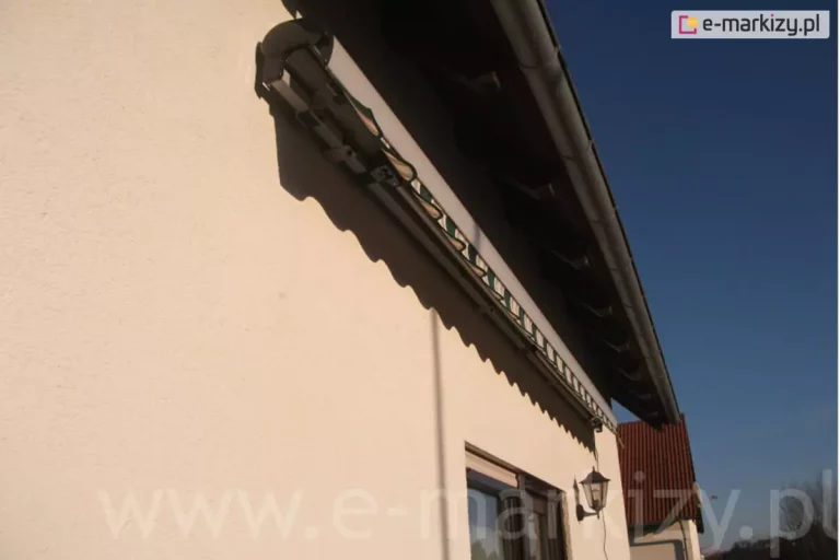 Zdjęcie przedstawia fragment elewacji budynku jednorodzinnego z zamontowaną markizą tarasową palladio aluprof. Markiza jest w stanie złożonym, zwinięta pod aluminiową kasetą, która osłania rurę nawojową z tkaniną od góry i z przodu przed działaniem czynników atmosferycznych. Widoczna tkanina markizowa ma wzór w pasy o kolorystyce zielono-beżowej z elementami bieli, zakończona jest ozdobną falbaną o falistym brzegu, która zwisa swobodnie z belki przedniej dosuniętej do kasety. Mechanizm został przytwierdzony do jasnej, tynkowanej ściany budynku, która ma delikatną strukturę ziarnistą. Konstrukcja markizy jest umiejscowiona bezpośrednio pod okapem dachu, gdzie widoczna jest podbitka dachowa wykonana z ciemnobrązowego drewna oraz metalowa rynna odprowadzająca wodę. Poniżej markizy znajduje się wnęka okienna oraz klasyczna lampa zewnętrzna w formie czarnego lampionu zamontowana na ścianie. W tle po prawej stronie widać fragment sąsiedniego budynku z dachem pokrytym czerwoną blachodachówką oraz bezchmurne, błękitne niebo, co sugeruje słoneczną pogodę i duży kąt padania światła, rzucający wyraźny cień markizy na elewację.