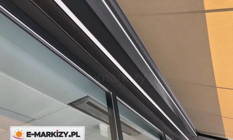 Zdjęcie prezentuje detal konstrukcyjny, jakim jest kompaktowa kaseta systemu markiza Moderno, zamontowana na elewacji budynku. Aluminiowa obudowa kasety w kolorze antracytowym posiada nowoczesny, kanciasty kształt o matowym wykończeniu, a w jej dolnej części widoczna jest precyzyjnie dopasowana listwa frontowa, która po złożeniu szczelnie domyka mechanizm. Z kasety wyprowadzone jest poszycie wykonane z wysokiej jakości tkaniny akrylowej w kolorze piaskowego beżu, która jest równomiernie napięta i wykazuje gęsty, techniczny splot. Konstrukcja jest zainstalowana bezpośrednio nad dużym przeszkleniem, w którym odbija się jasna podbitka dachu lub sufit pomieszczenia. Minimalistyczny design kasety sprawia, że dyskretnie integruje się ona z architekturą obiektu, chroniąc jednocześnie zwinięty materiał przed niekorzystnymi warunkami atmosferycznymi.