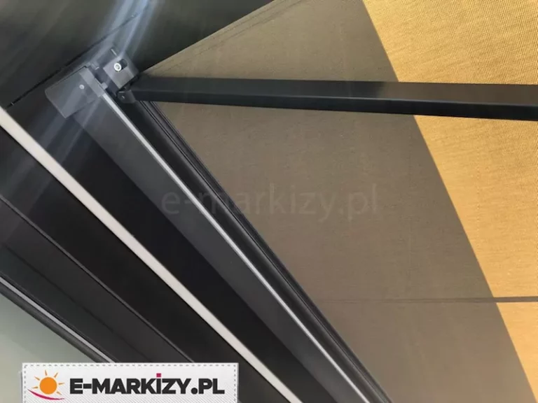 Zdjęcie prezentuje szczegół techniczny konstrukcji, jaką posiada markiza Moderno, skupiając się na systemie mocowania i prowadzenia ramion. Na pierwszym planie widoczny jest solidny, aluminiowy profil nośny w kolorze ciemnografitowym lub czarnym, do którego za pomocą metalowego wspornika zamontowane jest przegubowe ramię markizy. Konstrukcja ramienia charakteryzuje się prostą, nowoczesną linią i matowym wykończeniem. Powyżej stelaża rozciąga się gładkie poszycie materiałowe w odcieniu stonowanego beżu lub jasnego brązu, które jest napięte i stabilnie osadzone w profilu. Na tkaninie widoczna jest wyraźna granica światłocienia, co podkreśla gęsty splot materiału o wysokich właściwościach przeciwsłonecznych. Całość mechanizmu została zaprojektowana w sposób minimalistyczny, z widocznymi elementami montażowymi, takimi jak śruby imbusowe, co ułatwia precyzyjną regulację kąta nachylenia zadaszenia.
