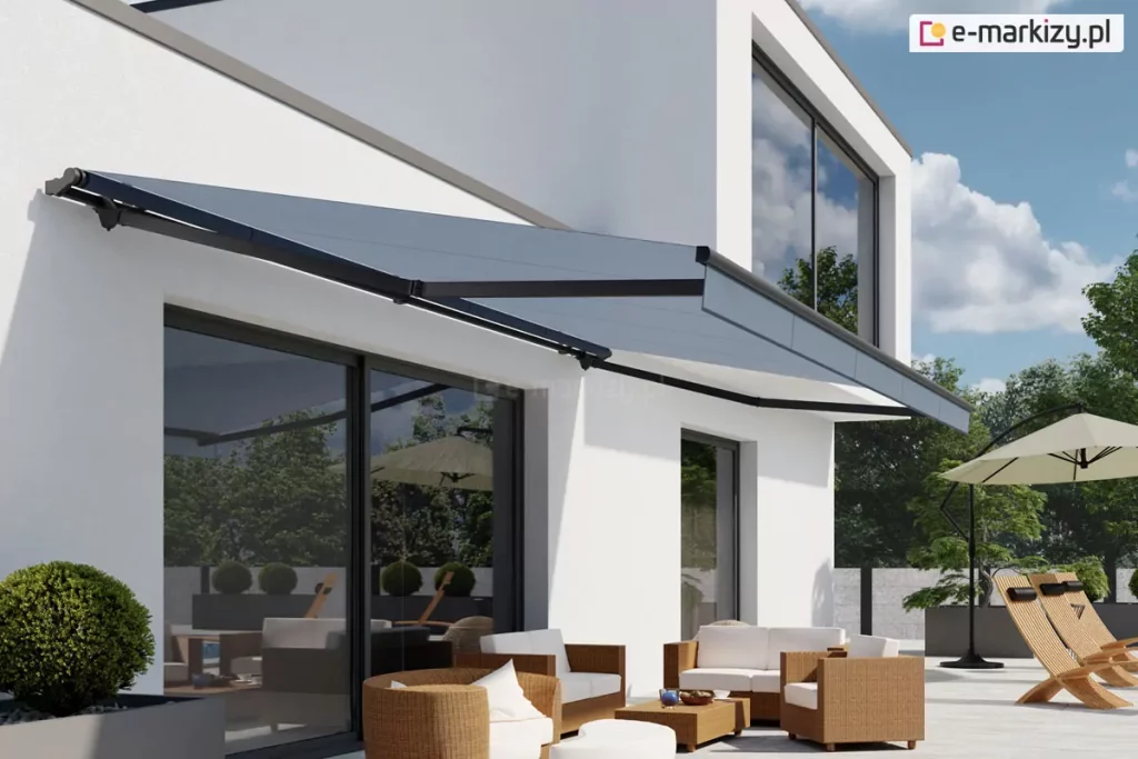 Nowoczesna szara markiza Australia zamontowana na białej, gładkiej elewacji piętrowego budynku mieszkalnego o minimalistycznej architekturze. Poszycie markizy w kolorze grafitowym jest w pełni rozwinięte i napięte na ciemnych, niemal czarnych ramionach przegubowych, rzucając wyraźny cień na taras oraz duże przeszklenia parteru. Konstrukcja markizy Australia charakteryzuje się brakiem pełnej kasety, co eksponuje jej techniczne detale montażowe przytwierdzone powyżej linii okien tarasowych. Pod markizą znajduje się zaaranżowana strefa wypoczynkowa z jasnobrązowymi meblami technorattanowymi z białymi poduszkami, ustawionymi na jasnoszarych płytach tarasowych. W tle widoczny jest ogród z zielonymi drzewami, krzewami formowanymi w kule oraz dodatkowe wyposażenie ogrodowe w postaci beżowego parasola bocznego i drewnianych leżaków o ergonomicznym kształcie. Całość kompozycji przedstawia produkt w pełnym słońcu na tle bezchmurnego, błękitnego nieba, podkreślając funkcję ochronną markizy Australia przed intensywnym promieniowaniem słonecznym.