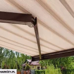 Zbliżenie na spód konstrukcji, którą posiada markiza Presto, prezentujące wytrzymałe, aluminiowe ramię przegubowe w kolorze ciemnego brązu. Zdjęcie ukazuje detal techniczny przegubu z widocznym potrójnym łańcuchem stalowym, który zapewnia stabilność i płynność ruchu podczas rozkładania materiału. Poszycie markizy wykonane jest z tkaniny w poziome pasy w tonacji beżu i jasnego brązu, która jest równomiernie napięta nad ramionami. W dolnej części kadru widoczna jest brązowa belka frontowa, do której przymocowane jest ramię, a w tle rozpościera się widok na zielony ogród z drzewami liściastymi oraz fragment sąsiedniego domu jednorodzinnego z żółtą elewacją i spadzistym dachem.