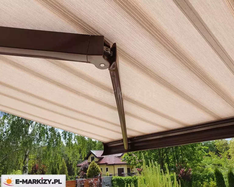 Zbliżenie na spód konstrukcji, którą posiada markiza Presto, prezentujące wytrzymałe, aluminiowe ramię przegubowe w kolorze ciemnego brązu. Zdjęcie ukazuje detal techniczny przegubu z widocznym potrójnym łańcuchem stalowym, który zapewnia stabilność i płynność ruchu podczas rozkładania materiału. Poszycie markizy wykonane jest z tkaniny w poziome pasy w tonacji beżu i jasnego brązu, która jest równomiernie napięta nad ramionami. W dolnej części kadru widoczna jest brązowa belka frontowa, do której przymocowane jest ramię, a w tle rozpościera się widok na zielony ogród z drzewami liściastymi oraz fragment sąsiedniego domu jednorodzinnego z żółtą elewacją i spadzistym dachem.